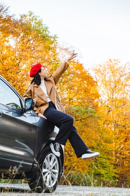 Una mujer linda con un atuendo elegante se sienta en el capó de un automóvil con la mano levantada y apuntando hacia el cielo