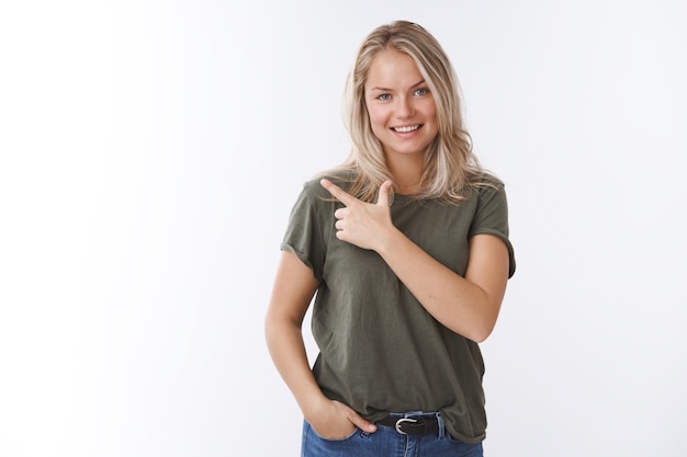 Mujer linda confiada invitando al hombre agarrar café apuntando en la esquina superior izquierda o detrás con el dedo índice sosteniendo la mano en el bolsillo y sonriendo a la cámara dando la bienvenida a una presentación amistosa sobre una pared blanca