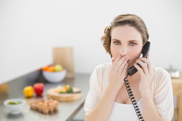 Mujer linda sorprendida llamando en su cocina con un teléfono