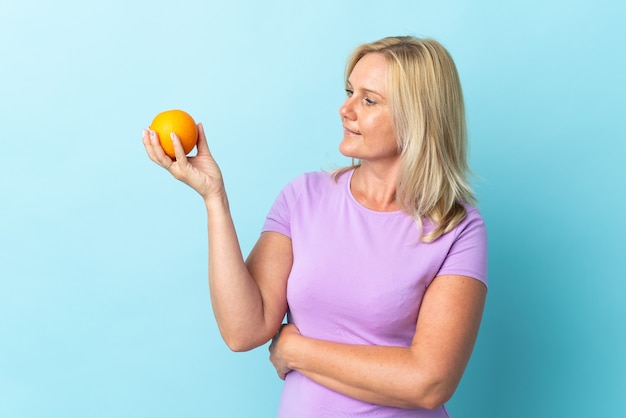 Mujer lituana de mediana edad aislada sobre pared azul sosteniendo una naranja