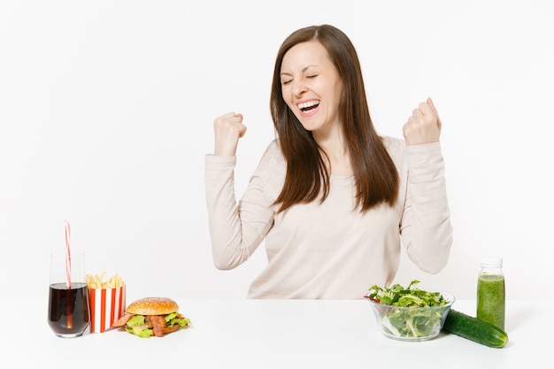 Mujer llena de alegría en la mesa con batidos de desintoxicación verde, ensalada en un tazón de vidrio, pepino, hamburguesa, cola en botella aislada sobre fondo blanco. Nutrición adecuada, estilo de vida saludable, comida rápida, concepto de dieta