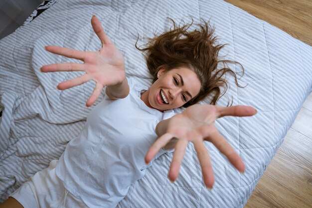 Mujer llena de alegría en pijama estirando los brazos después de despertarse acostado en una cama acogedora feliz sonriendo un ...