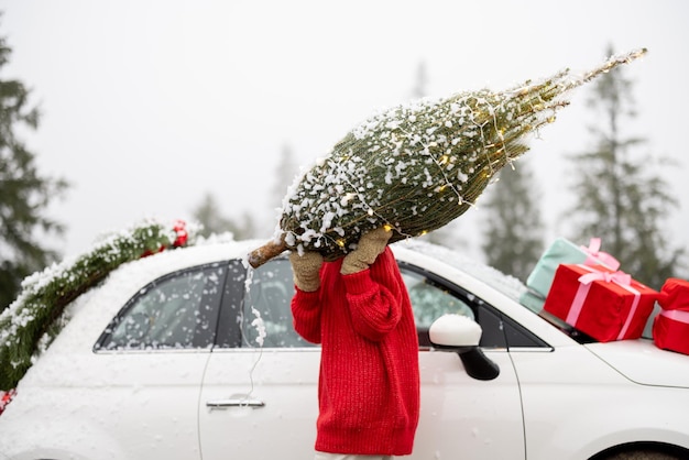 Mujer lleva un árbol de navidad envuelto cerca de un auto en las montañas