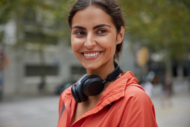 Mujer lleva auriculares anorak alrededor del cuello sonríe positivamente posa en borrosa