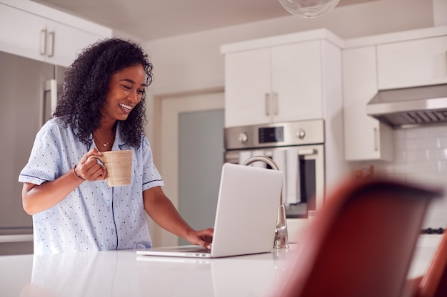 mujer, llevando, pijama, posición, en, cocina, trabajando, de, casa, en, computadora portátil