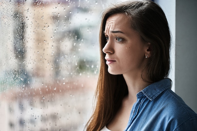 Foto mujer llorando con lágrimas en los ojos que sufre conmoción emocional, pérdida, pena, problemas de la vida y una relación de ruptura junto a la ventana con gotas de lluvia. hembra recibió malas noticias