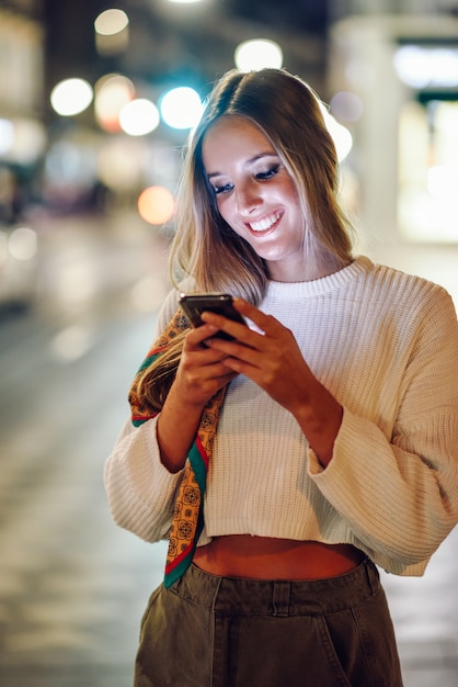 Mujer con las luces urbanas defocused de la ciudad que miran su smartphone.