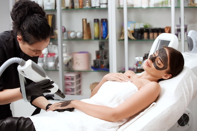 Mujer de lujo en spa con máscara de carbono en la cara y cosmetóloga trabajando en su brazo