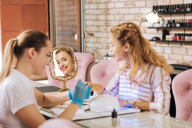 Mujer madura alegre curar las uñas bajo la lámpara UV y sonriendo al espejo