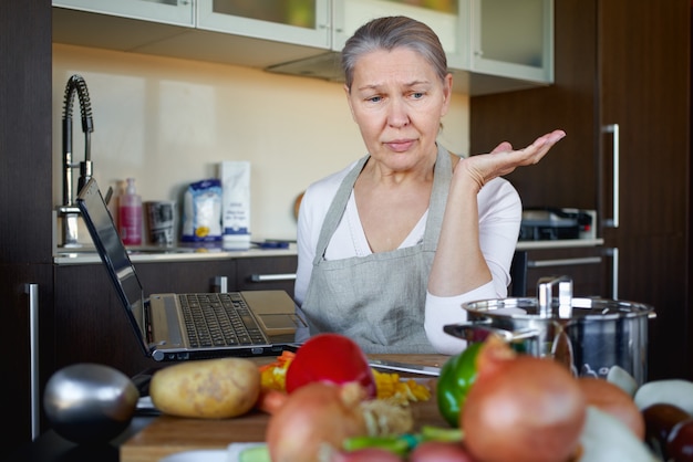 Mujer madura angustiada en la cocina prepara comida y usa laptop