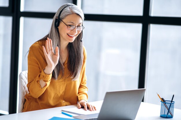 Una mujer madura asiática de cabellos grises sonriente está hablando en línea usa una computadora portátil para reuniones virtuales en línea saludando a colegas mientras videoconferencia concepto de enseñanza a distancia de comunicación en línea