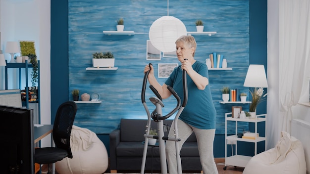 Mujer madura con bicicleta estática para hacer ejercicio físico en casa. Persona jubilada entrenando los músculos de las piernas con bicicleta paso a paso electrónica, haciendo ejercicios de fitness con máquina de cardio.