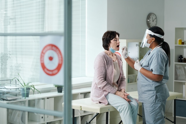 Mujer madura con la boca abierta sentada delante del médico en ropa de trabajo protectora probándola para covid con hisopo oral en consultorio médico