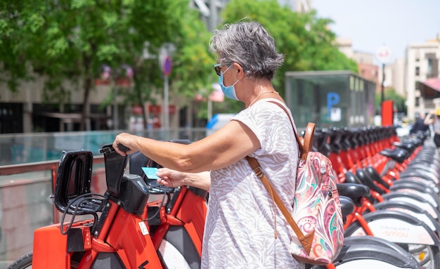 Mujer madura de cabello gris lista para alquilar una bicicleta eléctrica para un recorrido por la ciudad con una tarjeta de crédito para el pago Anciana activa con mochila con máscara