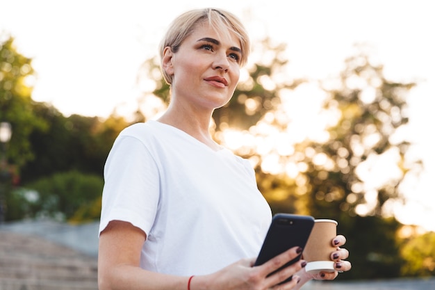 Mujer madura con cabello rubio vistiendo ropa casual sosteniendo un teléfono celular y una taza de papel con café, mientras está de pie en las escaleras de la ciudad