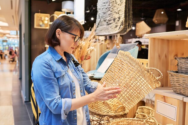 Mujer madura compradora eligiendo cestas de mimbre interiores en la tienda de decoración del hogar