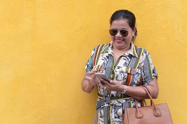 Foto mujer madura consultando un smathphone mientras se apoya contra una pared.