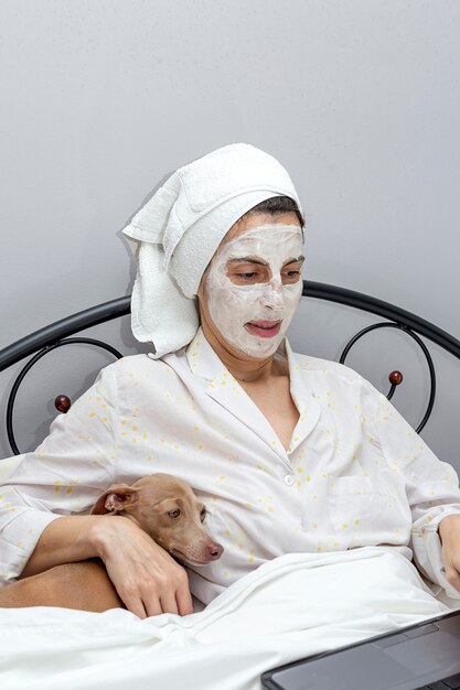 Mujer madura cuidando su rostro con una mascarilla en la cama con perro