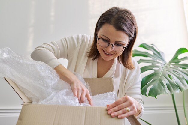 Mujer madura desempaqueta la caja de cartón en la mesa en casa, en la oficina. Servicio de envío postal internacional, auto entrega de mercadería, carga. La mujer recibió compras compradas en tiendas en línea