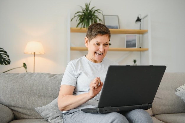 Una mujer madura emocionada mirando la pantalla de una laptop leyendo buenas noticias en un mensaje celebrando la victoria de la lotería en línea regocijándose del éxito mujer mayor feliz sentada en el sofá en casa usando una computadora