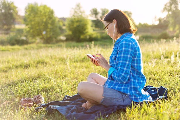 Mujer madura escucha música, un audiolibro en auriculares, se relaja en la naturaleza. Puesta de sol de fondo, paisajes rústicos, prado verde, espacio de copia