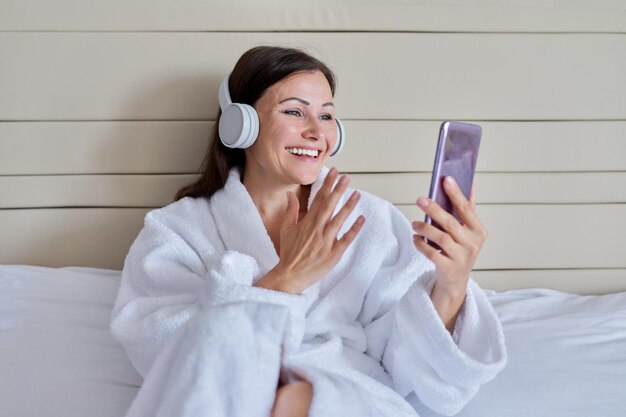 Mujer madura feliz en albornoz blanco y auriculares con teléfono inteligente sentado en la cama