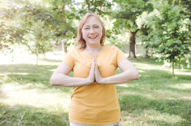 Una mujer madura feliz con una camiseta amarilla haciendo yoga en el parque. Estilo de vida activo