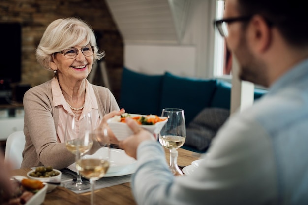 Mujer madura feliz comiendo con su hijo adulto y pasándole comida en la mesa del comedor