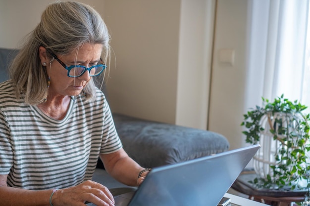 Mujer madura con gafas en casa usando una computadora portátil en el sofá