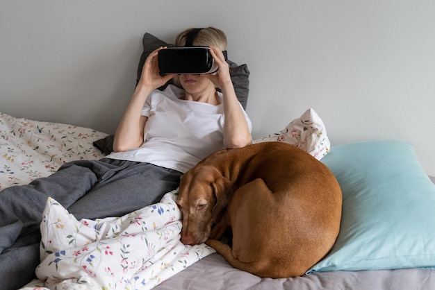 Mujer madura con gafas VR acostada en una cama sin hacer con mascotas juega y trata de evitar problemas de vida