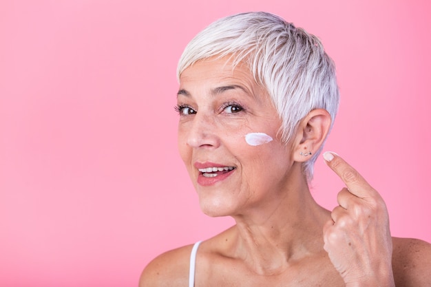 Mujer madura hermosa que sostiene el tarro de crema hidratante y que mira la cámara. Mujer mayor feliz que sostiene una botella de loción antienvejecedora aislada sobre fondo rosado. Tratamiento anti envejecimiento y belleza.