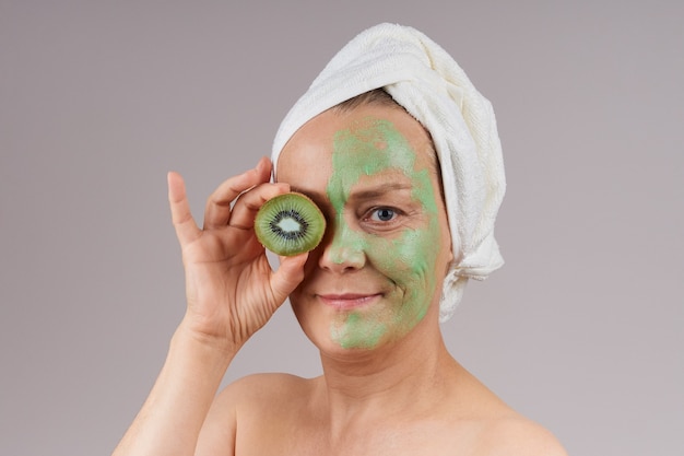 Mujer madura con hombros descubiertos, toalla blanca en la cabeza, mascarilla de fruta verde aplicada en el rostro, kiwi cerró los ojos. Concepto de cuidado de la piel facial. sobre la pared gris.