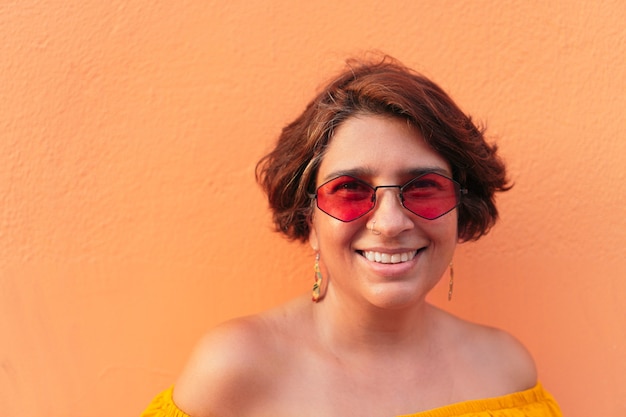 Mujer madura latina con blusa amarilla y gafas de sol sobre fondo naranja.