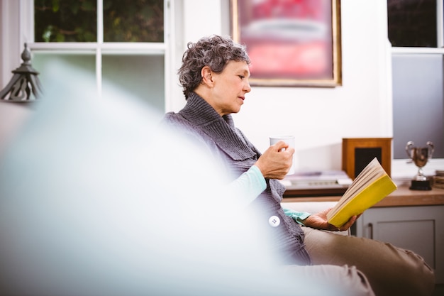 Mujer madura leyendo el libro y sosteniendo la taza de café mientras está sentado en el sofá
