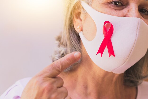 Mujer madura con máscara pandémica y cinta rosa aislada en blanco. Imagen del mes de concientización sobre el cáncer de mama.