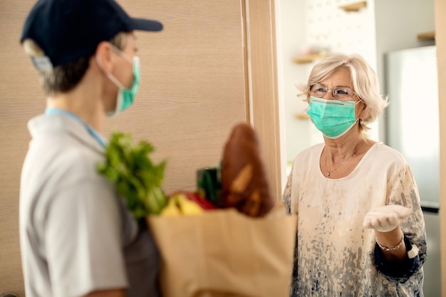 Mujer madura con mascarilla hablando con un mensajero que le está entregando comestibles en casa