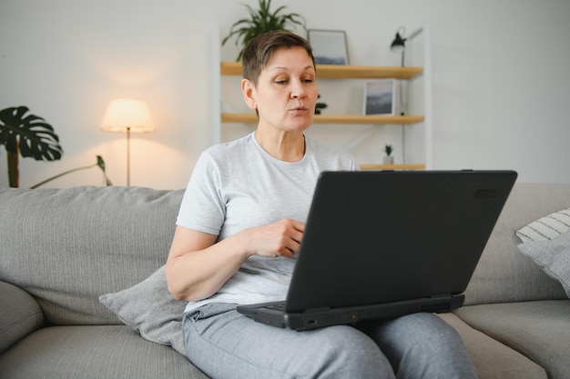 Mujer madura mayor que usa aplicaciones inalámbricas para portátiles navegando por Internet sentada en un sofá sonriendo abuela de mediana edad trabajando a distancia en la computadora navegando por la web comunicándose en línea mirando la pantalla en casa