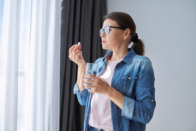 Mujer madura de mediana edad en ropa casual en casa con pastilla y vaso de agua dulce. Dolores de cabeza, depresión, medicamentos, suplementos, vitaminas, sedantes, menopausia