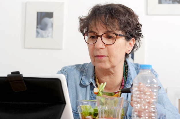 Mujer madura mirando tableta mientras come una ensalada