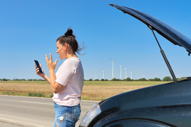 Mujer madura molesta conductora llamando por teléfono móvil para ayudar a una mujer cerca de un auto roto con el capó levantado en la carretera en el campo