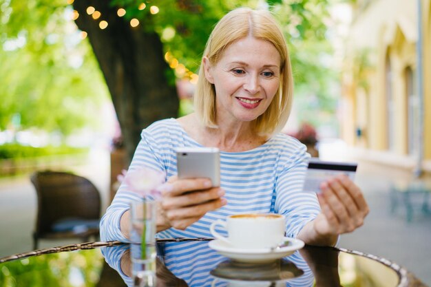 Mujer madura pagando con tarjeta bancaria usando un teléfono inteligente relajándose sola en un café de la calle