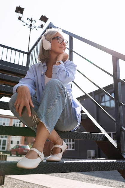 Foto una mujer madura de pelo gris vestida de verano escucha música con auriculares mientras camina por el