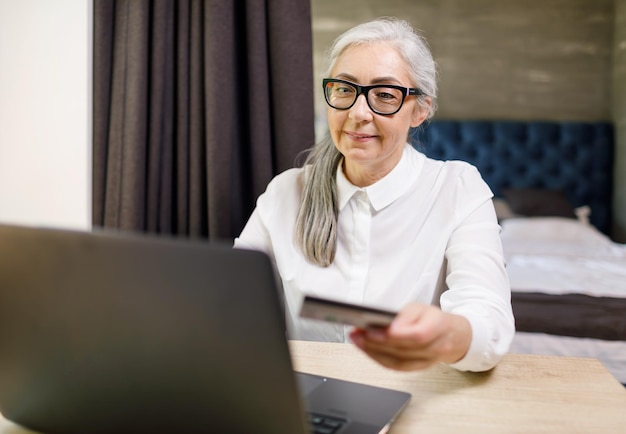 Mujer madura de pelo largo con gafas sentada con una laptop y comprando algo en Internet en su cómodo piso