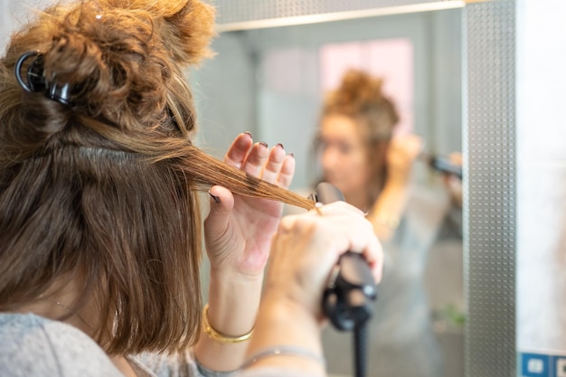 Mujer madura planchando su cabello frente al espejo en el baño de su apartamento