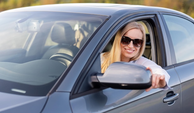 Mujer madura positiva con cabello rubio con gafas de sol y camisa azul ajustando el espejo lateral antes de conducir el automóvil Concepto de personas y transporte