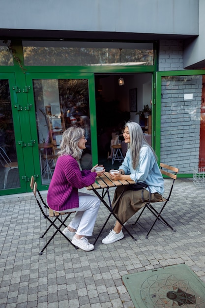 Mujer madura positiva y compañero asiático de pelo gris con café en la terraza de la cafetería al aire libre