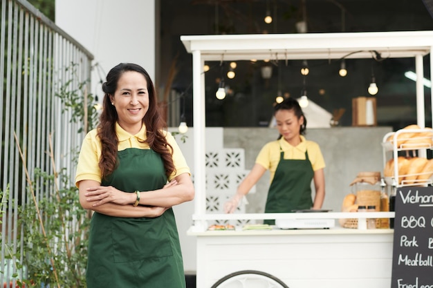 Mujer madura positiva en delantal de pie frente al carrito de comida cuando su hija está cocinando en segundo plano.