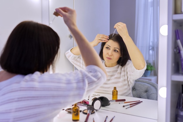 Mujer madura que trata el cuidado del cabello usando productos médicos cosméticos en botella con pipeta Mujer de mediana edad sentada en casa mirándose en el espejo Problema de pérdida de cabello preparaciones profesionales especiales
