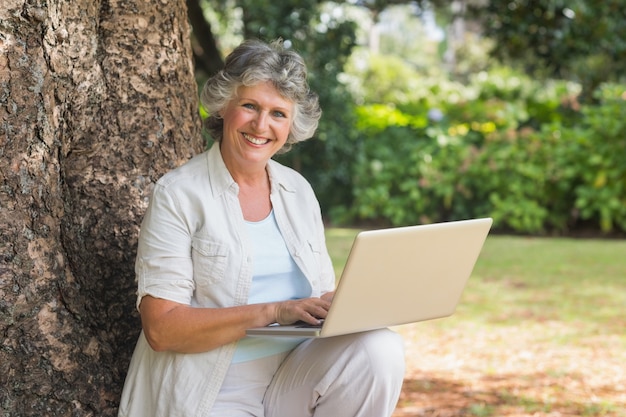 Mujer madura que usa una computadora portátil que se sienta en el tronco de árbol