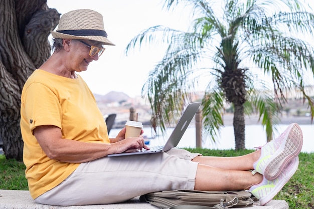 Mujer madura relajada sentada al aire libre usando una computadora portátil sosteniendo una taza de café